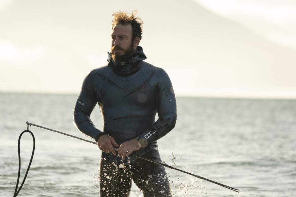 Freediver Grant Hogan walking out of the Pacific ocean at sunrise in a wetsuit holding a fishing spear